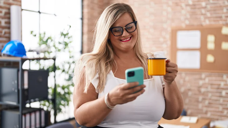 Mulher, em pé e com sobrepeso, sorri ao olhar a tela do celular