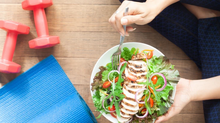 Mulher comendo salada e aparelhos de academia ao lado