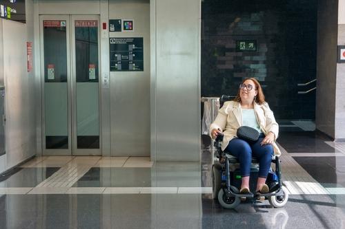 Mulher cadeirante aguarda em frente ao elevador com piso tátil em ambiente acessível e adaptado.