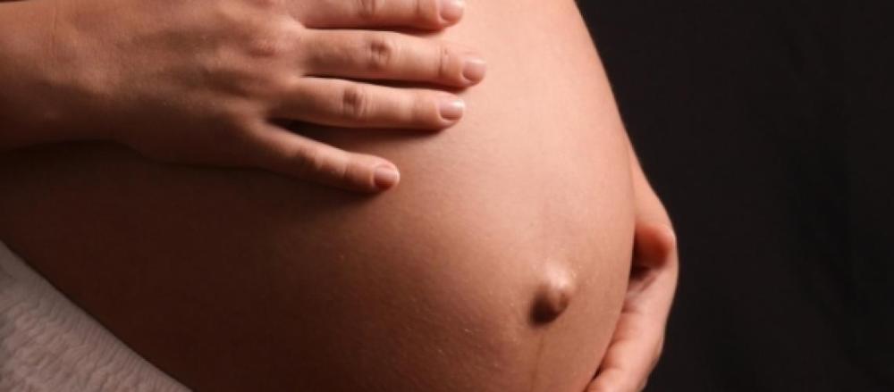 A imagem mostra um close do abdômen de uma pessoa grávida. Uma das mãos repousa suavemente sobre a parte superior da barriga, enquanto a outra apoia a lateral inferior. A cena destaca o formato arredondado da gestação e transmite uma sensação de cuidado, conexão e proteção.